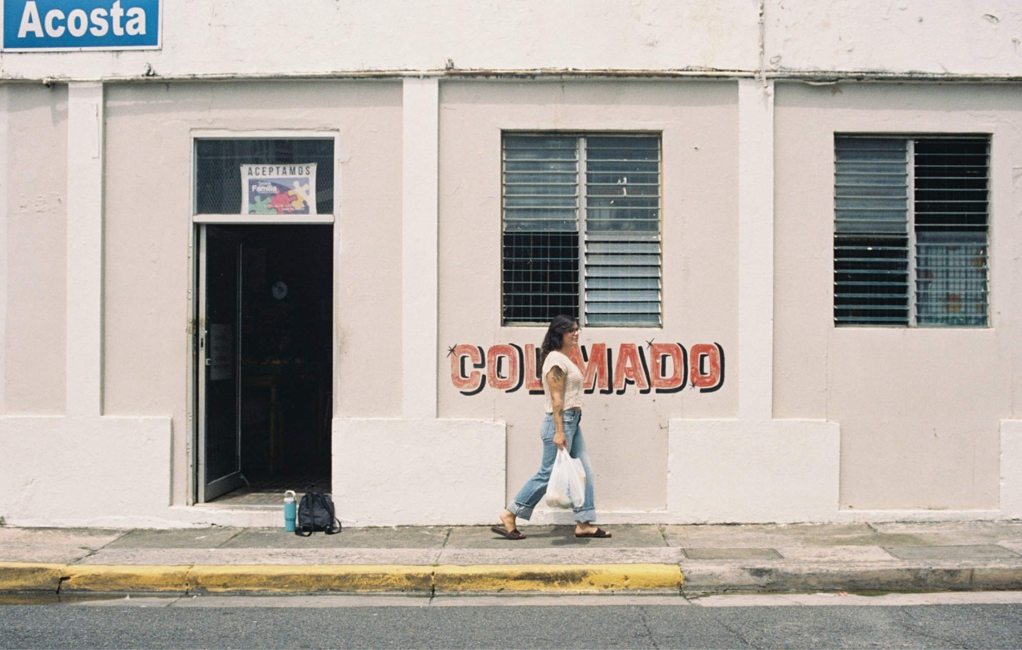 Person walking past a pale pink corner storefront with a colmado sign and turquoise window shutters.