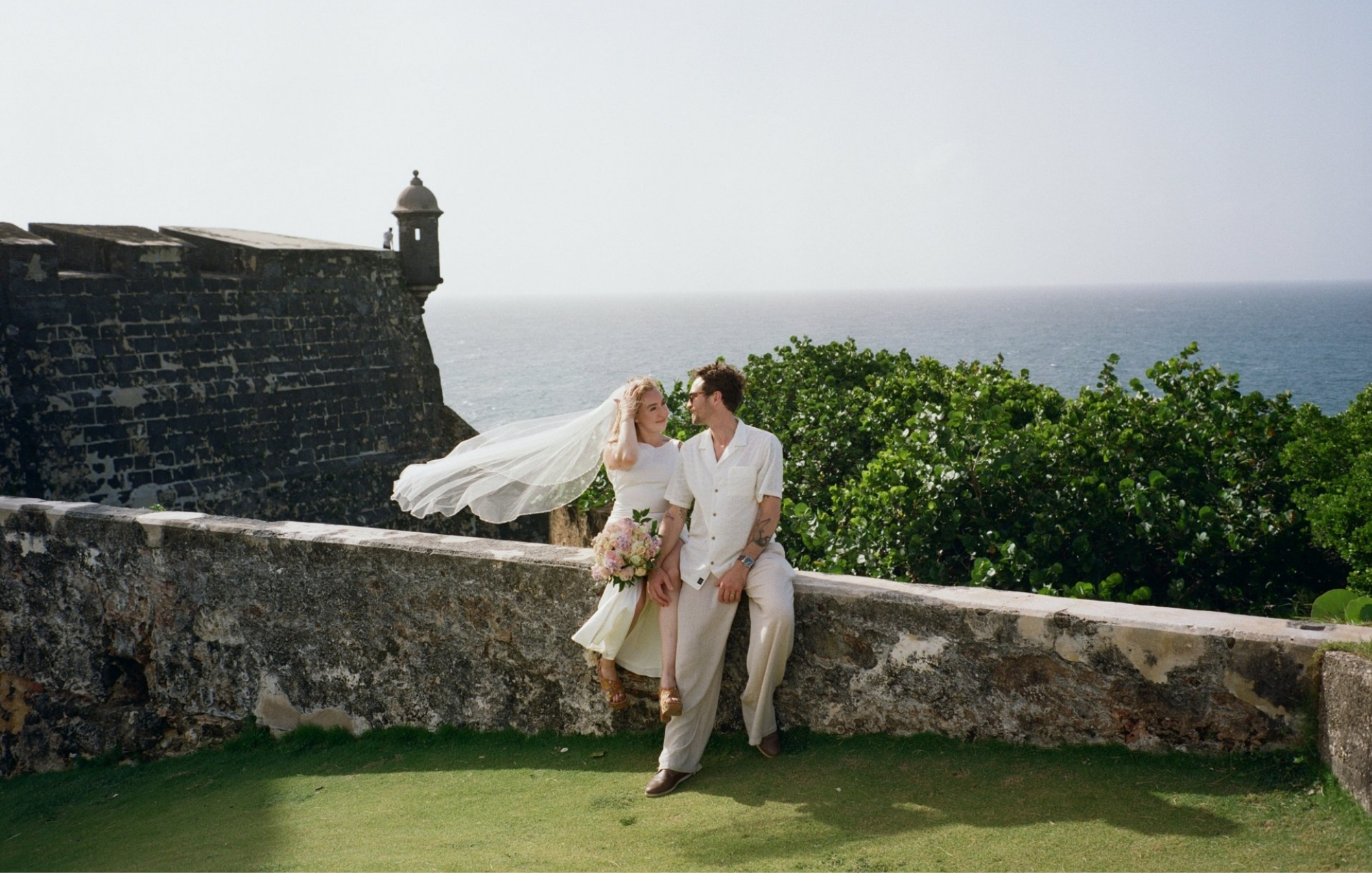 Wedding couple standing beside a historic stone fort overlooking the ocean, with the bride’s veil blowing in the breeze.