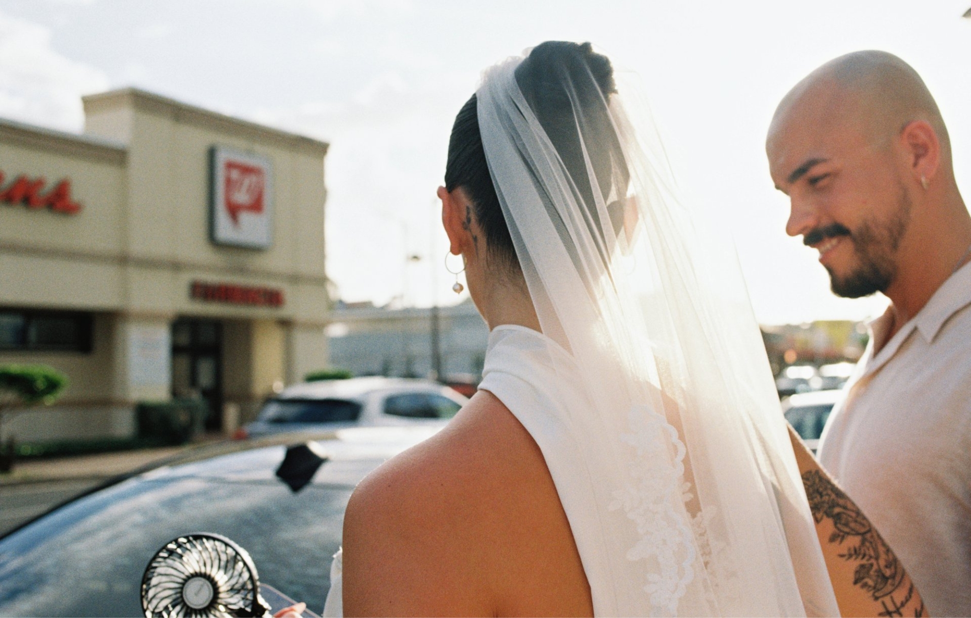 Bride and groom smiling beside a car outside a shopping center, with the bride’s veil in the sunlight